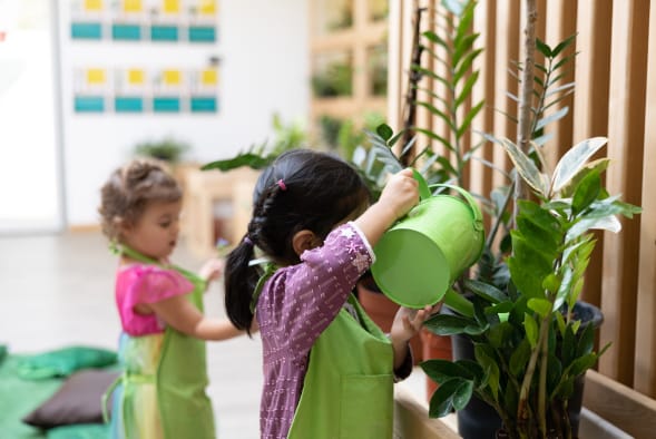 Two girls in green aprons are playing with plants in a classroom.